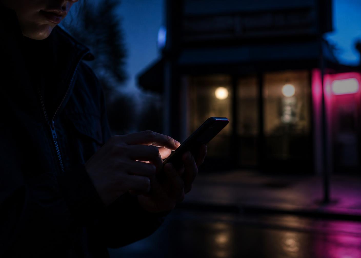 Pessoa buscando no celular à noite, em frente a uma loja de bairro iluminada